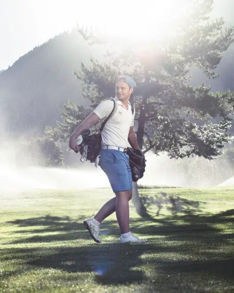 Mann mit Golftasche geht auf einem sonnigen Golfplatz mit Bäumen und Bergen im Hintergrund.