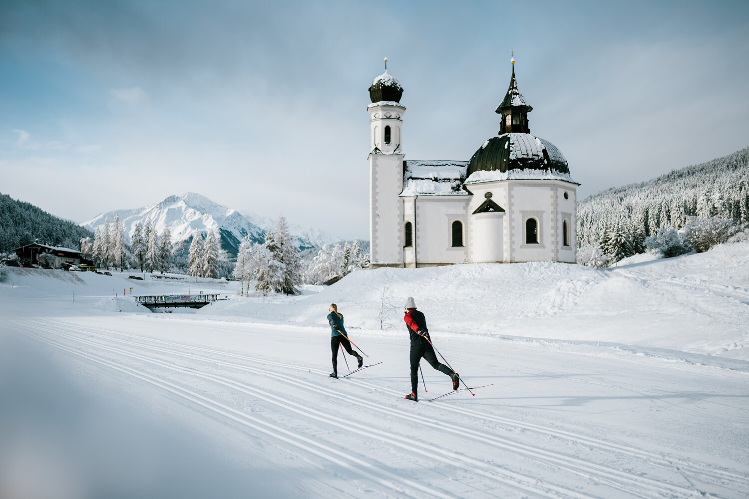 Zwei Personen laufen Langlaufski in der Nähe einer verschneiten Kirche in der malerischen Seefelder Winterberge.