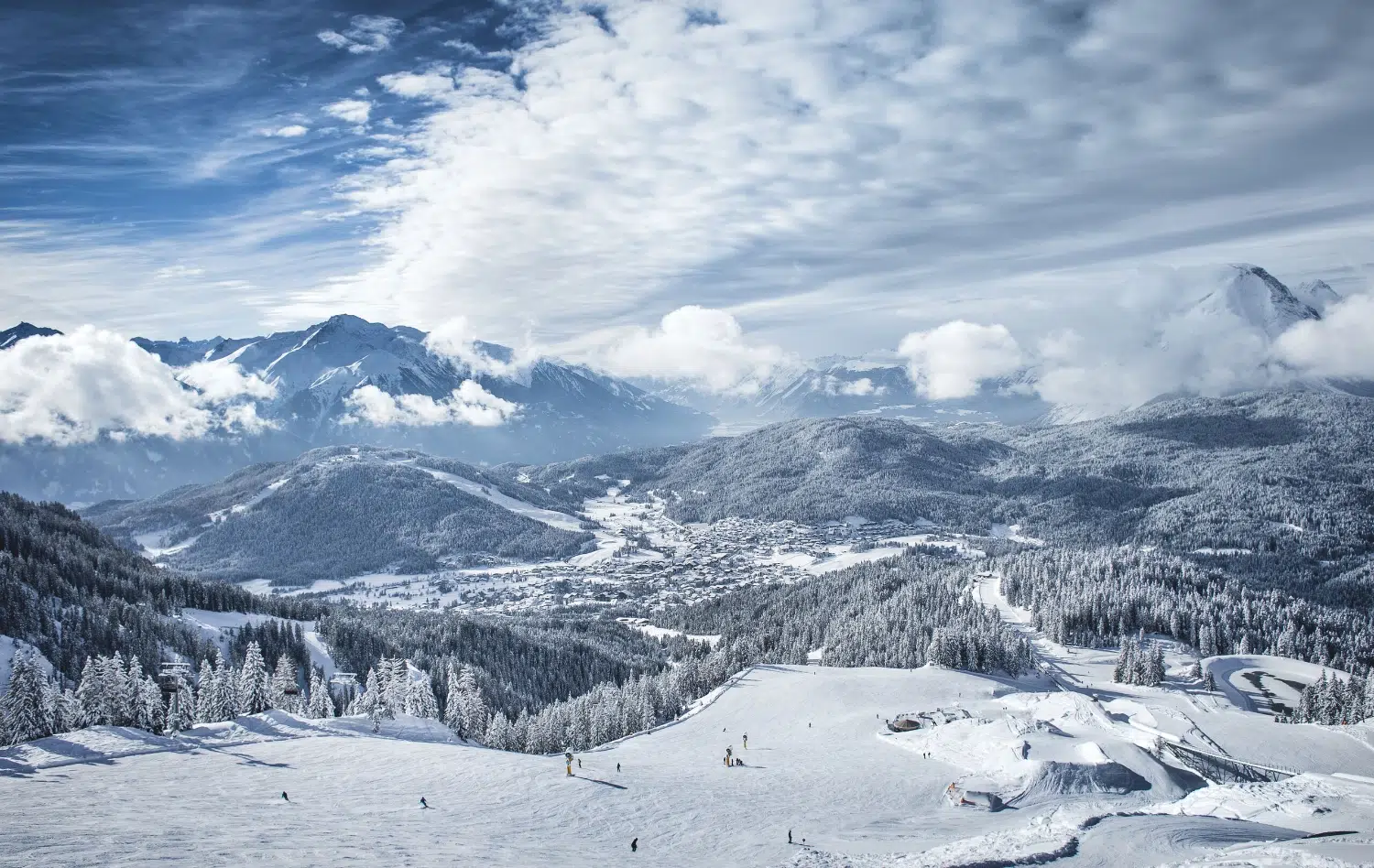 Schneebedeckte Berge und bewaldete Hügel mit einem Dorf im Tal unter einem bewölkten blauen Himmel.