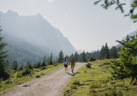 Zwei Personen gehen auf einem Pfad durch eine malerische, sonnenbeschienene Berglandschaft mit Bäumen.