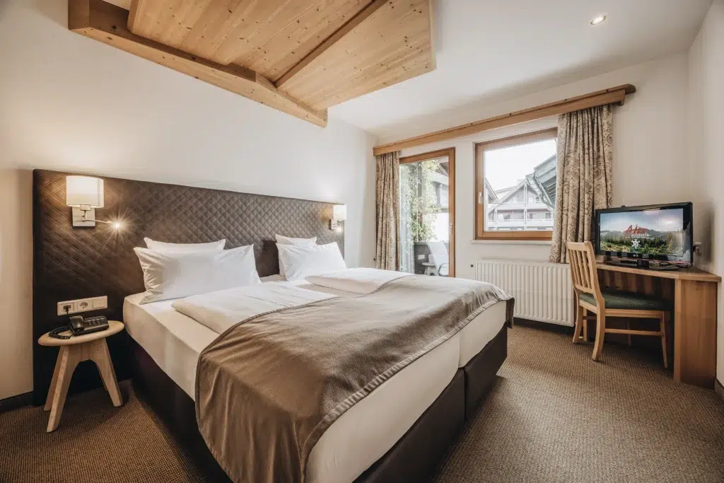 Gemütliches Zimmer in der Ferienwohnung Seefeld mit Doppelbett, Fernseher, Schreibtisch, Fenster und Holzakzenten an der Decke.