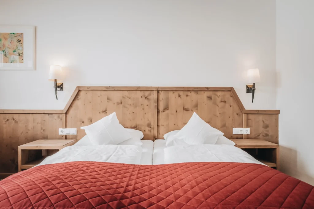 Minimalistisches Seefeld-Komfortzimmer mit zwei Einzelbetten, roter Steppdecke, Holzkopfteil und Wandlampen.