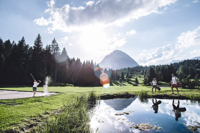 Drei Personen spielen Golf an einem Teich mit Bergen, Bäumen und dem 4-Sterne-Hotel Seefeld im Hintergrund.
