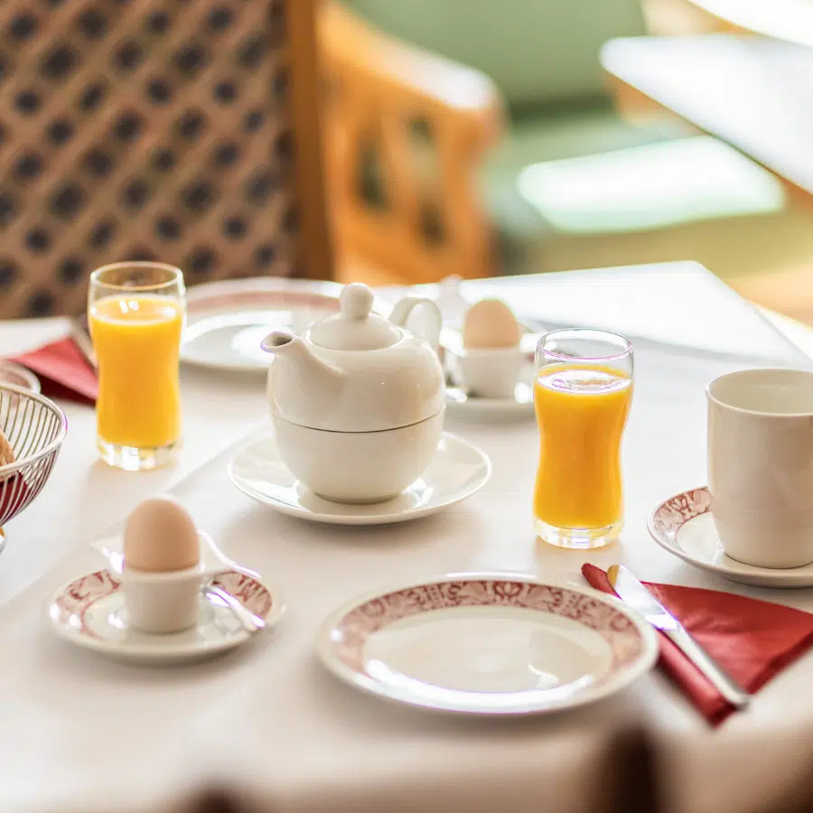 Frühstückstisch mit Eiern, Orangensaft, einer Teekanne, Tassen und Tellern auf einer weißen Tischdecke.