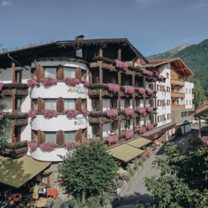 Alpenhotel mit Balkonen, die mit rosa Blumen gesäumt sind, Berge im Hintergrund.