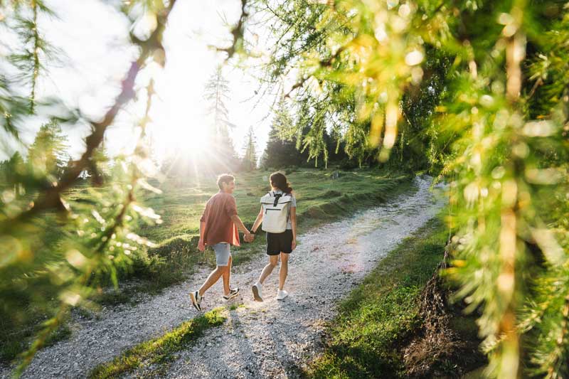 Zwei Menschen, die sich an den Händen halten und auf einem Waldweg im Sonnenlicht spazieren gehen.