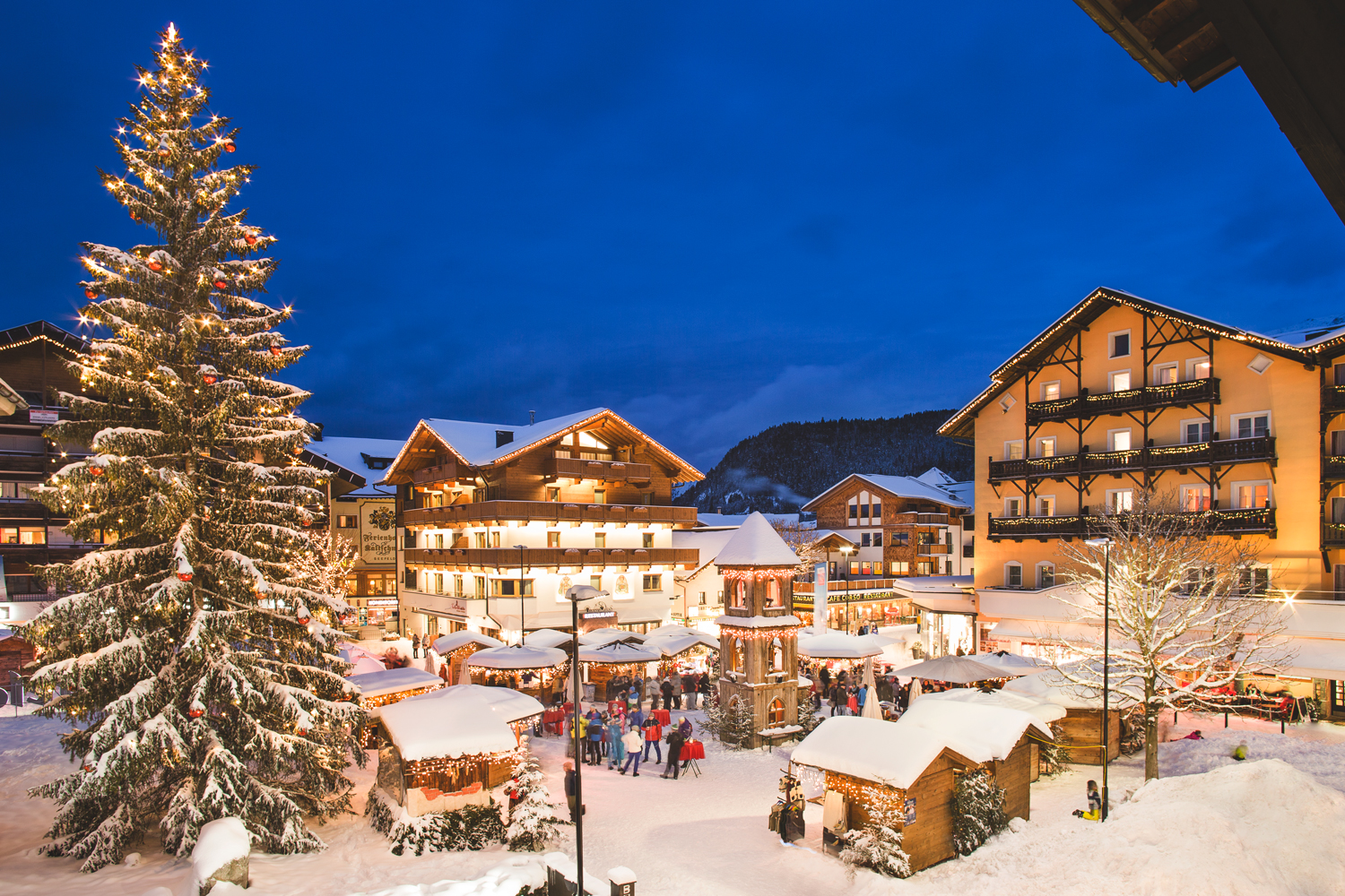 A snowy village square with a large lit Christmas tree and festive buildings at night.