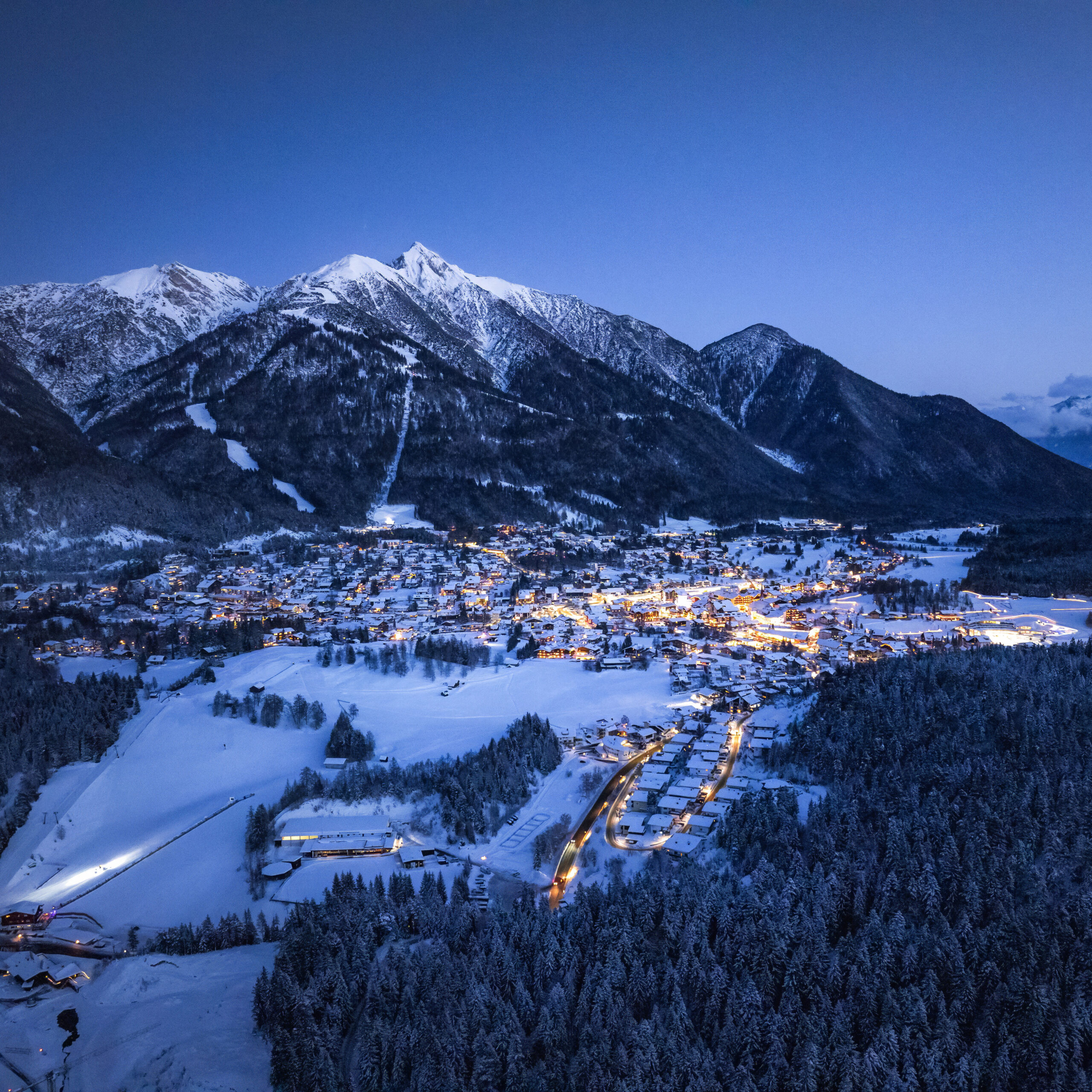 Snowy village at dusk with glowing lights, near a 4 star hotel Seefeld, amid mountains and trees.