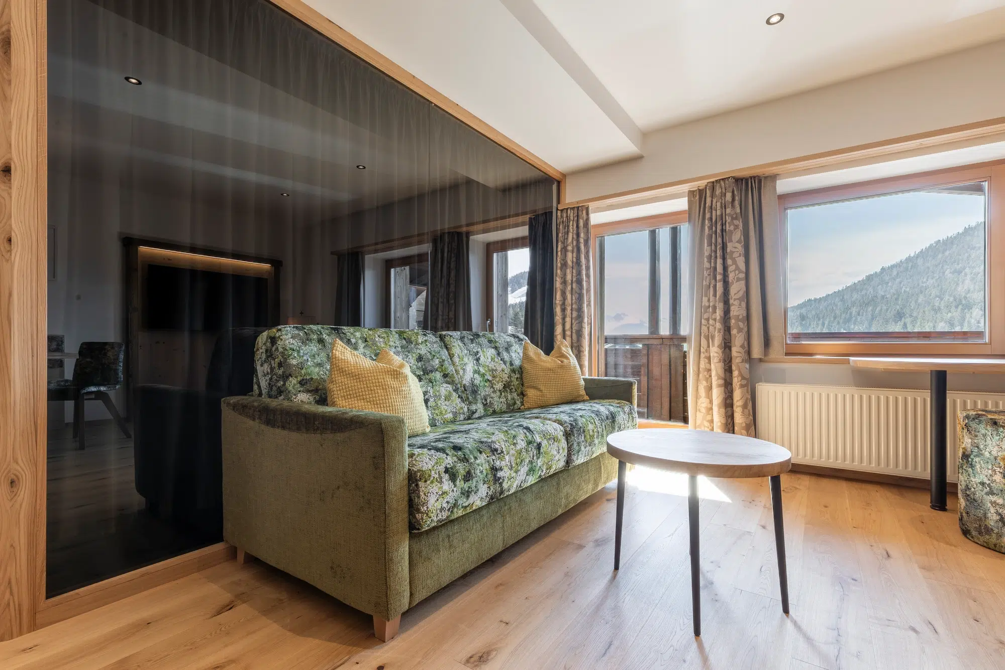 Grün gemustertes Sofa und runder Tisch in einem hellen Raum mit großen Fenstern und Bergblick.