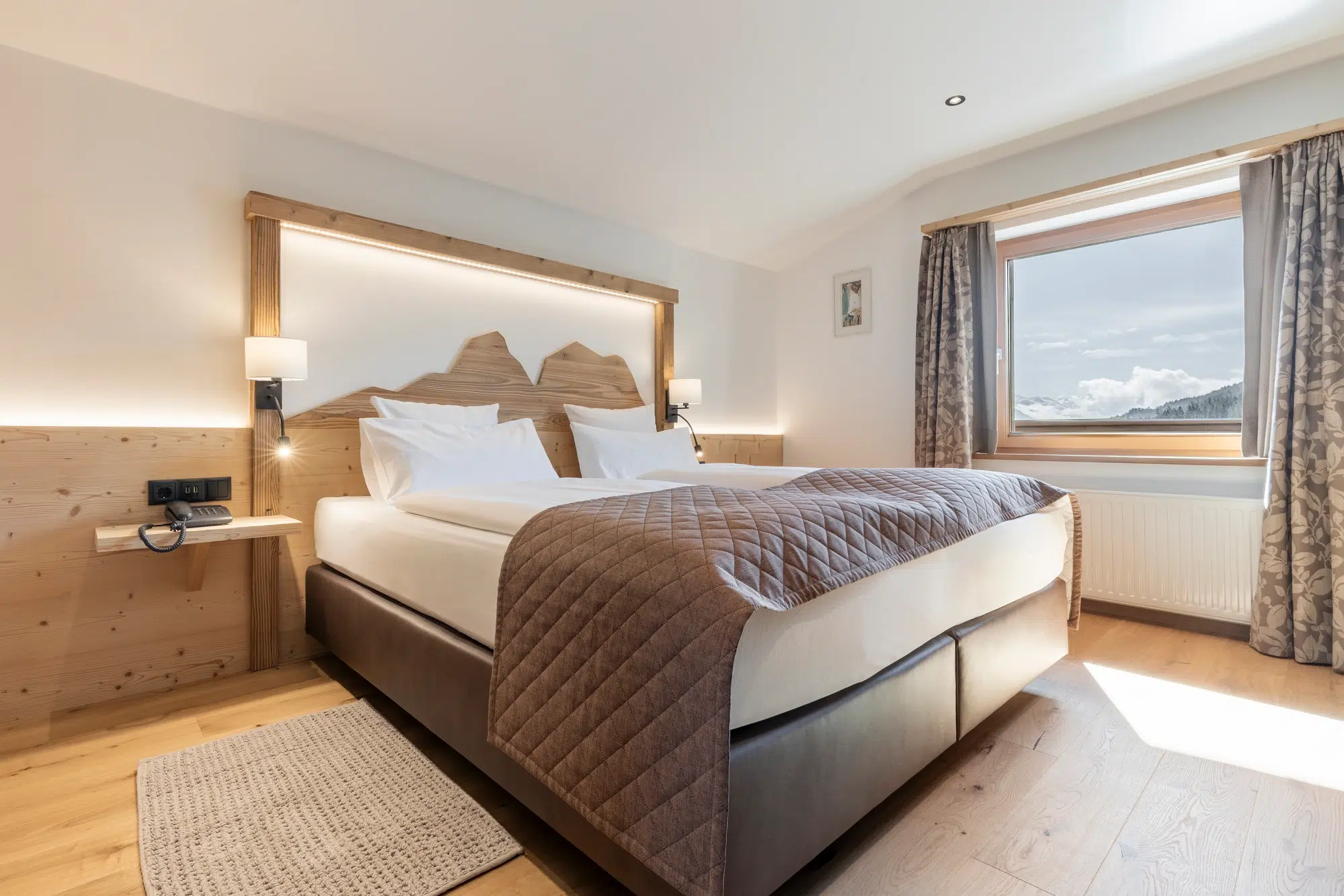Modernes Hotelzimmer mit großem Bett, Blick aus dem Fenster und einem Kopfteil aus Holz mit Bergmotiven.