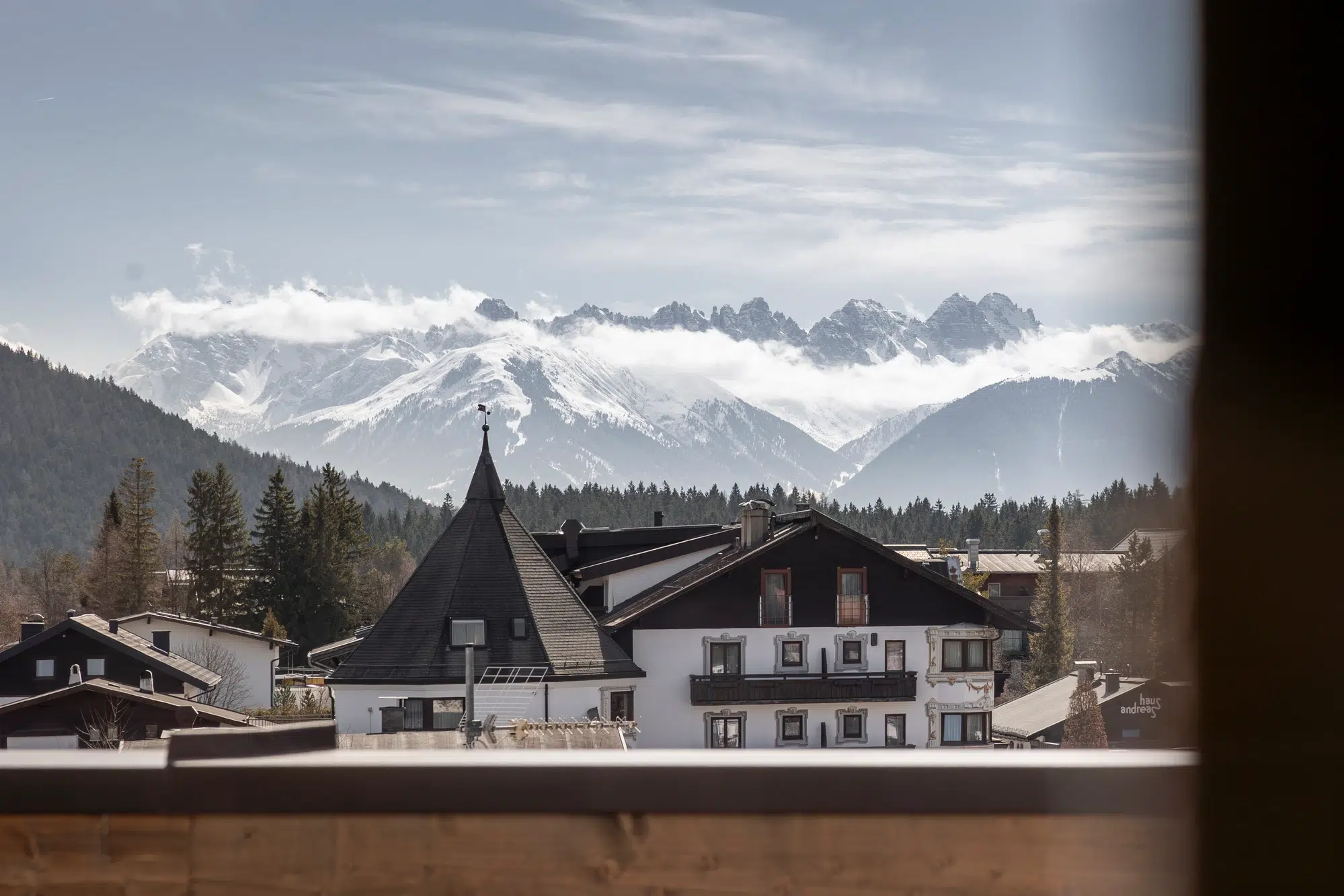 Schneebedeckte Berge und Tannenbäume hinter Gebäuden im alpinen Stil bei teilweise bewölktem Himmel.