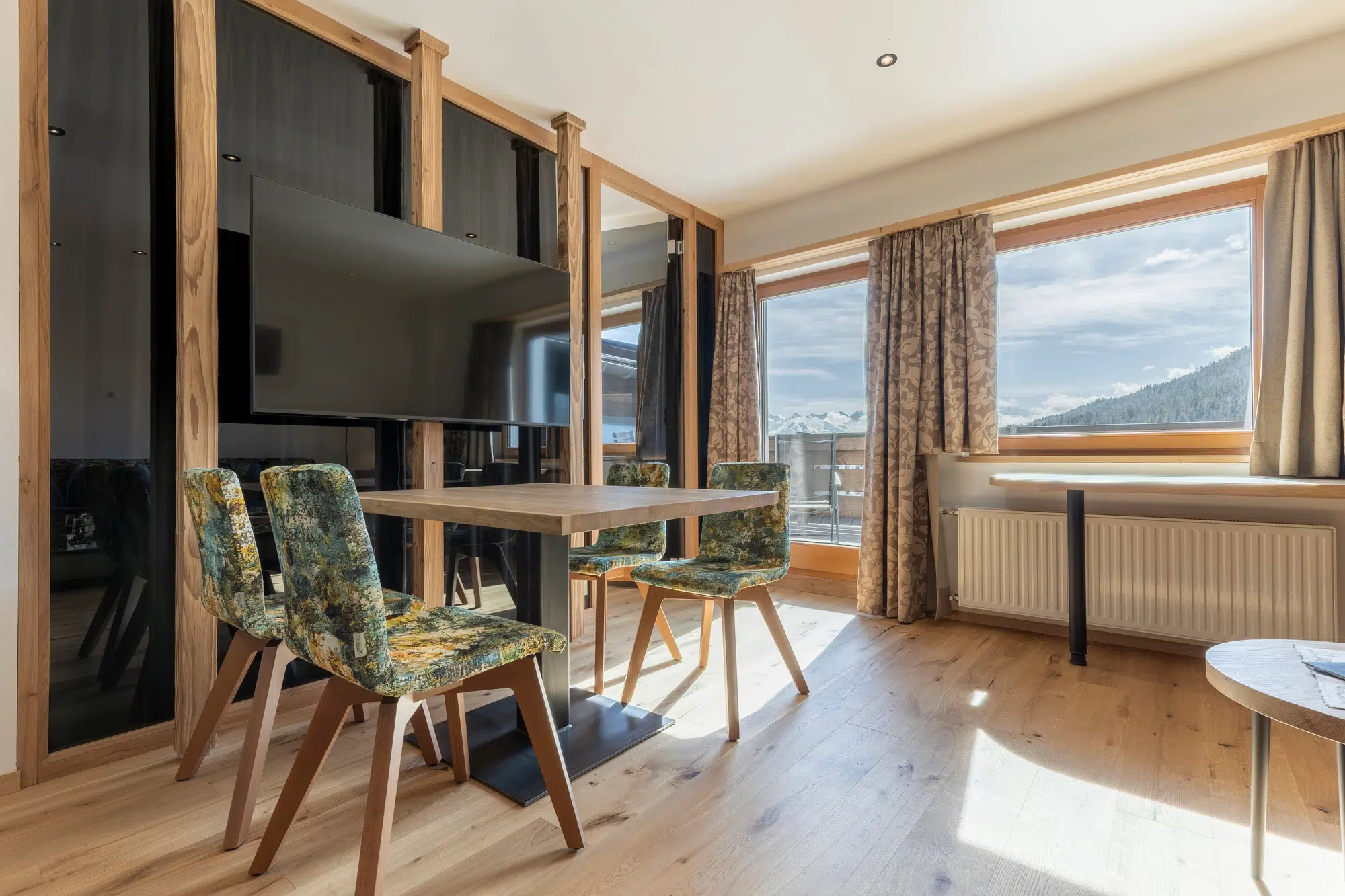 Sonniger Essbereich mit Holztisch, gemusterten Stühlen, TV und großen Fenstern mit Vorhängen.